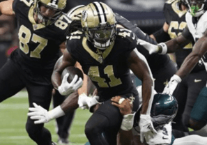 Football action scene with players in black and gold jerseys pushing forward against opponents in white and green. Intense, dynamic moment on the field.